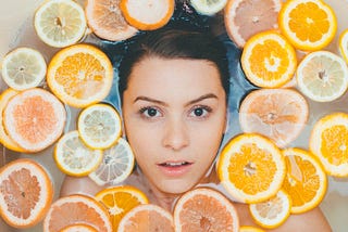 A make-up free woman looks out from a bathtub filled with sliced citrus fruit.