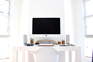 A neat white room with a picture of White Mac Monitor with symmetrically placed speakers, a white keyboard and a brown pot with green plant on one side and 2 brown desktop decorations of similar size on the other side of Monitor. All of this on a white table with a white ergonomic chair. A perfect symmetrical shot of this home office.