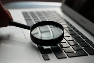 A person holding a magnifying glass close to a keyboard on a macbook computer.