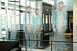 An artistic installation resembling the Star Trek transporter effect, featuring life-sized human figures seemingly formed from colorful beads suspended on vertical strings. The figures appear to be materializing or dematerializing in mid-air, capturing the iconic look of Star Trek’s transporter beam. The scene is set in a modern, spacious indoor area with natural light, glass walls, and minimalistic furnishings.