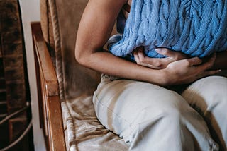 A woman sitting on a chair, bent forward while grabbing her stomach indicating stomach pain.