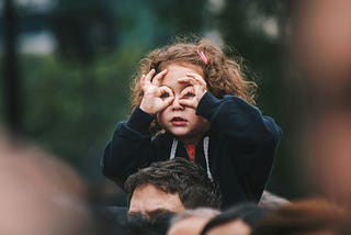 Curly-haired young girl sitting atop a man’s shoulders making round goggles over her eyes with her fingers — image being used by writer to depict looking for life’s purpose