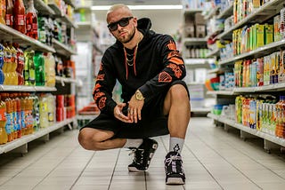 A tough guy wearing sunglasses and a hoodie crouching down in the drinks aisle of the supermarket
