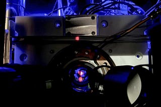 Close-up of a scientific instrument with wires and a circular metallic component, illuminated by blue light, featuring a small red dot at the center.