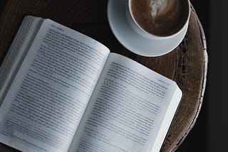 A novel is placed on a wooden table along with a cup of coffee.