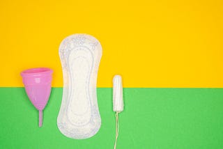 A close-up photo showing a tampon, a sanitary pad, and a menstrual cup placed side by side on a clean, white background, representing various menstrual hygiene products.