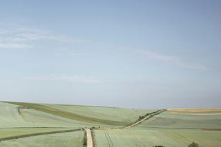 A dusty road going through green fields with a blue sky. People are hiking their way on the road