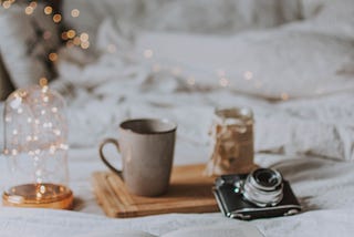 Book, open-faced on a bed with various items. A glittery light creates a magical feel next to a coffee mug on a tray and vintage camera.