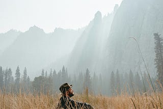 Hunter-like guy with a hat sitting in a wild landscape with pinetrees in the background and mountains.