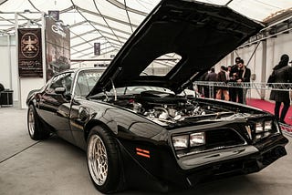 A black Pontiac Trans Am with an open hood in an exhibition hall.