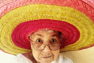 A close up image of an old lady wearing wire rimmed glasses. She is wearing an enormous red and yellow striped sombrero.