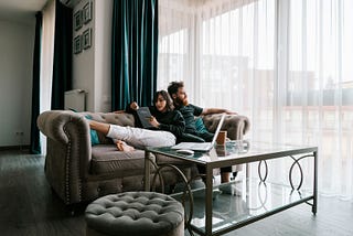A woman reading a book and a man looking outside sitting together on a sofa