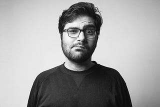 Black and white image of bearded man with glasses making a puzzled, wincing face