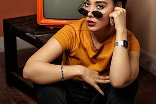 A young, attractive lady is sitting on the floor of a room facing the camera. She lowered her sunglasses on her knows so that we can see her eyes. She is wearing a yellow t-shirt and has a watch on her left arm and a silver link bracelet on her right arm. Behind her is a wooden television stand with a small TV screen on it.