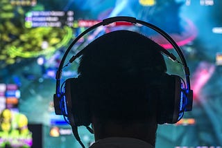 The man in the image is sitting in front of a computer screen and wearing headphones while playing a video game. The use of headphones highlights the importance of sound design in gaming, allowing players to fully appreciate the immersive and detailed audio experience that can greatly impact gameplay.