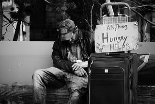 A dejected looking  man sitting with a suitcase and a sign lettered “Hungry Anything Helps” and “God Bless”