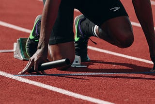 Relay racer at starting line, knee on the ground, toes set on blocks, holding a baton, ready to sprint. This image symbolizes the initial phase of market research, preparing businesses to launch forward with strategic insights.