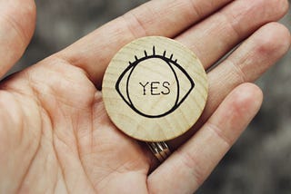 An open hand with a wooden disc displaying an eye with the word “yes” in the center.