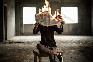 Man sitting on a stool inside an abandoned building holds a newspaper that is fire