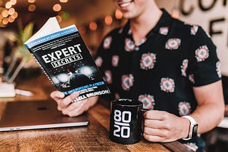 White guy with short sleeve shirt holding black mug with numbers on it reading a book about expert secrets