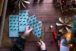 a pair of hands cut up some blue bags imprinted with a reindeer pattern for making paper Chrsitmas stars. Two other stars made of wrapping paper can be scene along with a glue stick and wreath.