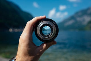 Photo of a lake in a mountain. In the foreground the photographer holds a camera lens. In the lens we can see the distant view magnified.