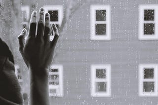 Hand on window, person looking out to building with several windows, on a rainy day