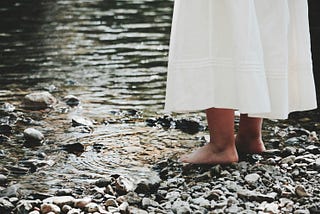 A girl stood barefoot on a river’s edge.