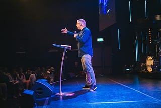 A man in casual dress addresses an audience from a stage