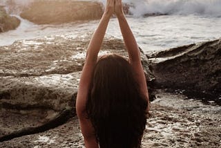 An image of a woman sitting on some rocks by the ocean. The waves are gently crashing into the rocks. The woman is wearing a white one piece swimsuit. She appears to be meditating. Her hands are clasped above her head and her hair is gently flowing down her back.