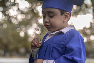 Child wearing graduation outfit blowing dandelion.