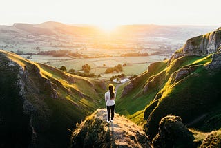 Woman standing on top of hill overlooking nature fields during sunset