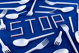 Bunch of single-use plastics spread out on a blue table, with the word “STOP” made from plastic straws, surrounded by many plastic spoons & forks.