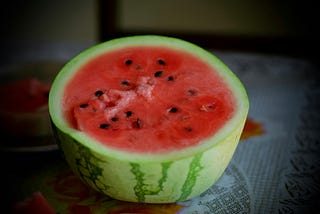 A picture of half of a watermelon, with its red center facing upwards on a tablecloth. The watermelon’s red center is also filled with dispersed black seeds.