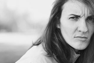 A black-and-white photo of a woman, with shoulder-length hair, looking over her shoulder and scowling at something in the distance.