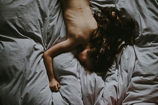 Naked woman with long brown hair, lying on her stomach on fluffy, gray bedding. Photo by Annie Spratt on Unsplash.