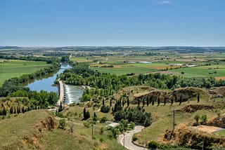 A photo of a landscape, with green fields, it’s a sunny day, a road zig-zags downhill and goes towards a bridge, that crosses the Duero river in Toro, Spain.