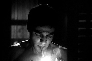 A black and white still of Sal Mineo as Lawrence Sherman in the 1965 film Who Killed Teddy Bear? The screen is dark, except for his face which is illuminated by a match he is using to light a cigarette in his mouth.
