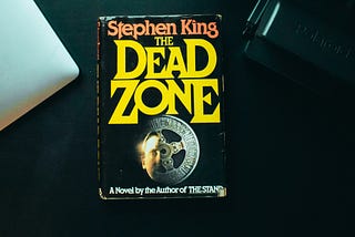 A book by Stephen King, Titled: The dead zone, on a background with a macbook laptop peaking out.