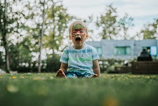 A young boy sitting in the grass and screaming with his mouth open.