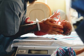 This image shows a newborn baby being held gently in a hospital setting right after birth. The baby appears to be fresh from delivery with a purplish hue, wet hair, and a hospital ID band around the wrist.