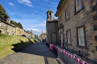 wide flagstone-paved path. To the left is a grassy bank with a terrace of stone houses at the top. To the left, a larger stone building with 4-paned windows and a clock tower. Iron railings run along the front of the building to the right. The railings are decorated with triangular flags with and “H” logo on them