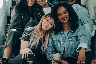 A group of seven women of different races and ethnicities sitting and smiling,  sitting on some steps.
