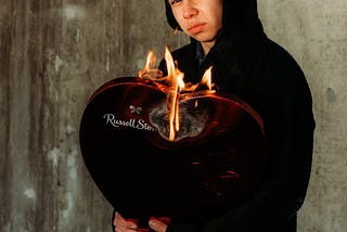 Hoodied man holding a heart-shaped candy box that’s on fire.