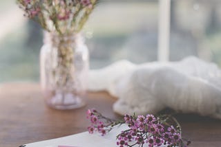A bouquet of purple delicate flowers in a mason jar on a wood desk. There is a notebook and a small stack of flowers are resting on it.