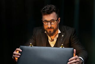 Man with a beard and wearing a suit and glasses looking into the camera while opening a briefcase in front of him. The contents of the briefcase, though we can’t see it, is glowing onto the man’s face