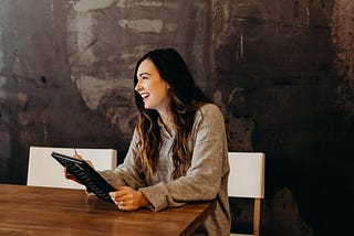 Woman smiling while sitting at a table and holding a tablet.