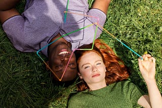 Two individuals laying on the grass with one person holding a thin colourful design sculpture