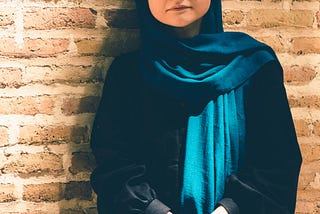 Woman standing against a wall wearing a deep blue Chador (long cloak, women in Iran wear that covers head and body leaving the face visible.)