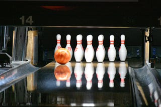 Orange bowling ball moving down an alley toward a ten pin setup.